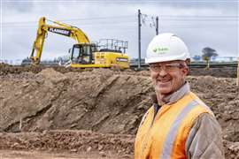 A man wearing an orange hi-vis vest and white hard hat on a construction site with an excavator in the background to his left